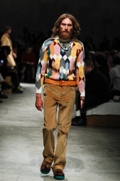 Prada Herbst/Winter 2017/18 Prada Herbst/Winter 2017/18 Bärtiger Mann mit langen Haaren, buntem Strickpullover und braunen Cordhosen.