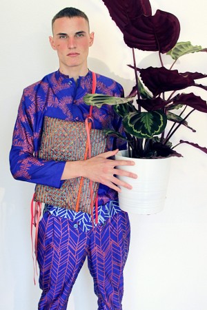 Männliches Model in violettem Outfit mit grafischem Muster. Hält Topfpflanze in der Hand.