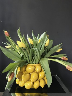 Foto/Styling: Nikolas Feireiss Gelber Tulpenstrauß vor schwarzem Hintergrund in runder mit Keramikvase, die mit Zitronen besetzt ist.