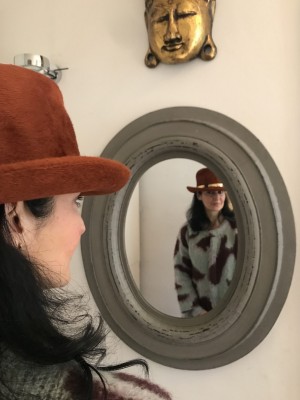 Frau mit Hut vor rundem Spiegel. Nadja Al-Chalabi